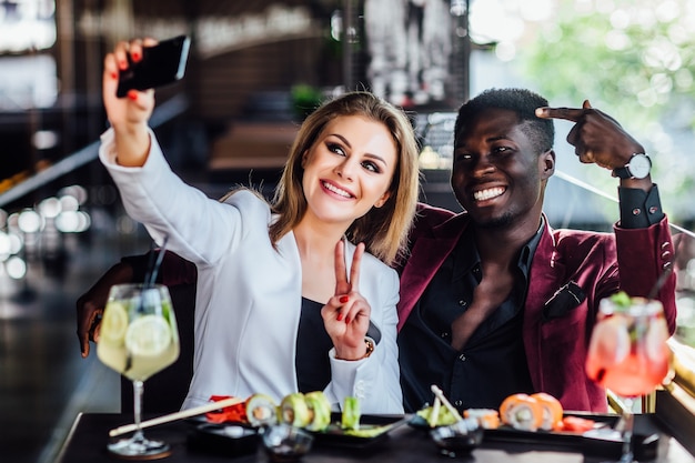 Hübsches blondes Mädchen, das ein Foto am Handy mit Sushi und Mojito macht. Chenese essen, Freundespaar.