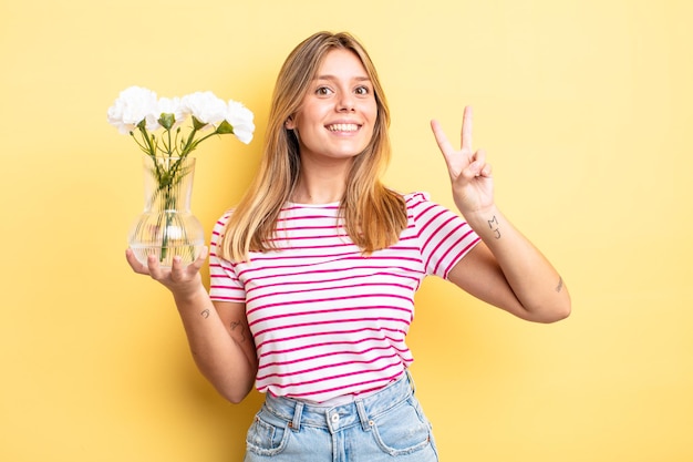 Hübsches blondes Mädchen, das freundlich lächelt und aussieht und Nummer zwei zeigt. dekoratives Blumenkonzept