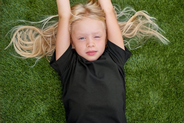 Hübsches blondes Mädchen des Nahaufnahmeporträts, das liegt und sich auf grünem Gras ausdehnt Mockup schwarzes T-Shirt Draufsicht