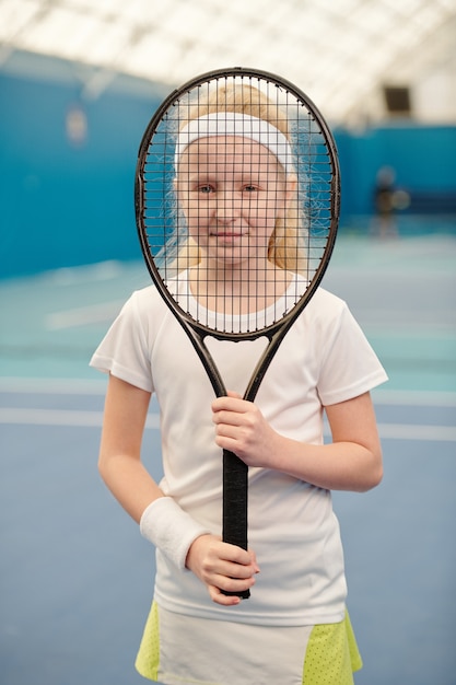 Hübsches blondes Mädchen in weißer Activewear, das Tennisschläger vor ihrem Gesicht hält und dich durch das Netz in der Stadionumgebung ansieht