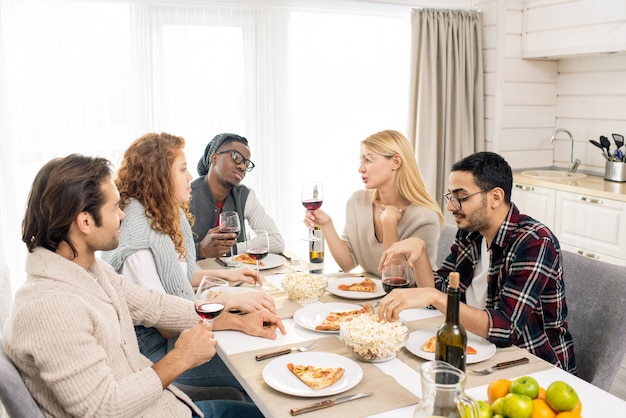 Hübsches blondes Mädchen mit Glas Wein, das unter ihren Freunden am servierten Tisch sitzt und feierlichen Toast bis zum Mittagessen ausspricht