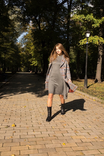 Hübsches blondes Modell mit langen Haaren in Strickkleid und warmem Mantel, das am sonnigen Tag im Park spazieren geht