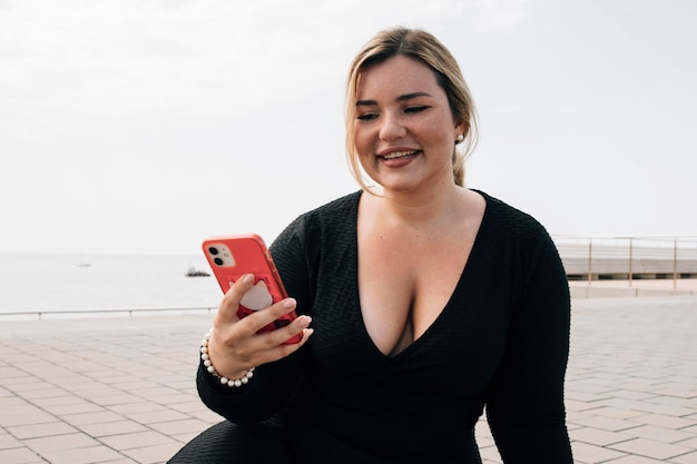 Hübsches blondes Plus-Size-Mädchen, das Nachrichten beantwortet oder auf ihr Handy vor dem Strand in Sportkleidung schaut