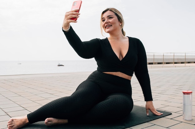 Hübsches blondes Plussize-Mädchen, das ein Selfie mit ihrem Handy vor dem Strand in Sportkleidung macht