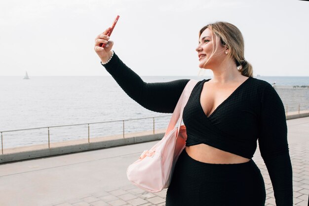 Hübsches blondes Plussize-Mädchen, das ein Selfie mit ihrem Handy vor dem Strand in Sportkleidung macht