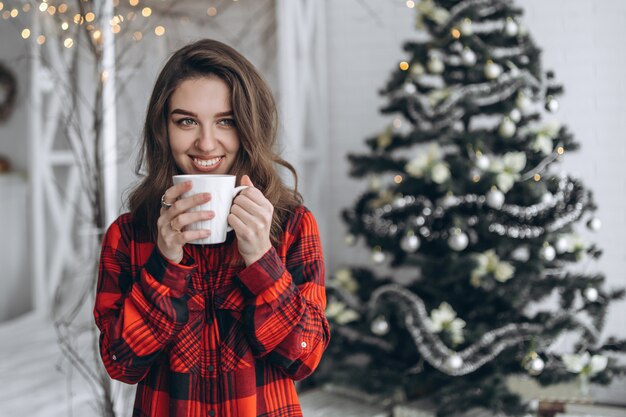 Hübsches brünettes Mädchen, das nahe Fenster steht und rotes Hemd und warme Socken mit Tasse Kaffee und Weihnachtslichtern dahinter trägt