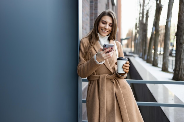 Hübsches brünettes Mädchen im braunen Mantelmantel mit Kaffeetasse mit Smartphone im Freien