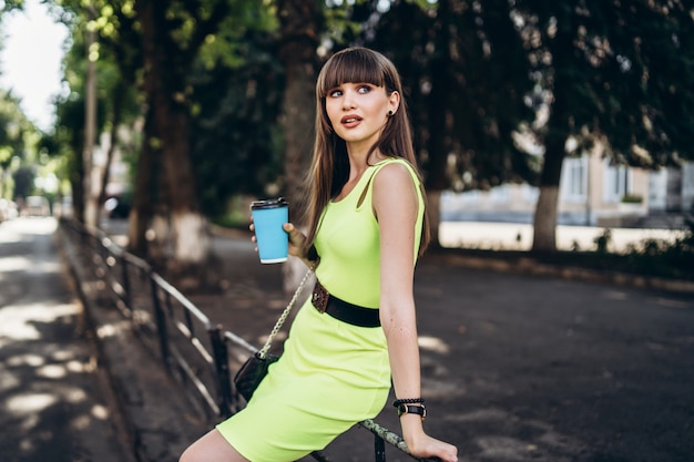 Hübsches brünettes Mädchen im grünen Kleid mit Tasse Kaffee, der draußen auf der Straße geht