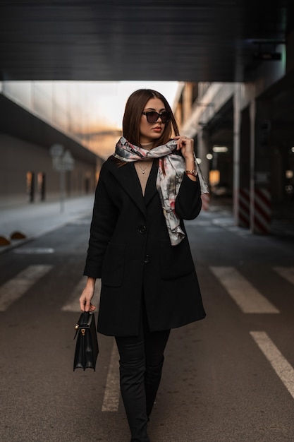 Hübsches Business-Modell der jungen Frau mit elegantem Schal auf dem Kopf in modischer Sonnenbrille in einem trendigen Mantel mit schwarzer Lederhandtasche steht auf der Straße in der Stadt. Sexy Mädchen, das draußen aufwirft. Moderne Dame.