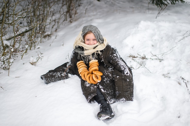 Hübsches Dorfmädchen steht im Schneewald mit getrockneten ringförmigen Brötchen und lächelt im Winter