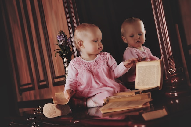 Hübsches einjähriges Mädchen im rosa Kleid mit einem alten Buch sitzt auf der Anrichte durch den Spiegel. Kind im Bibliotheksinnenraum. Konzept der Bildung und Kindererziehung. Hintergrund für Website oder Banner. Platz kopieren