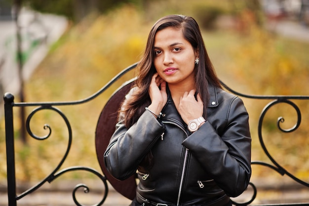 Hübsches indisches Mädchen in schwarzem Saree-Kleid und Lederjacke posierte im Freien auf der Herbststraße und saß auf einer Bank