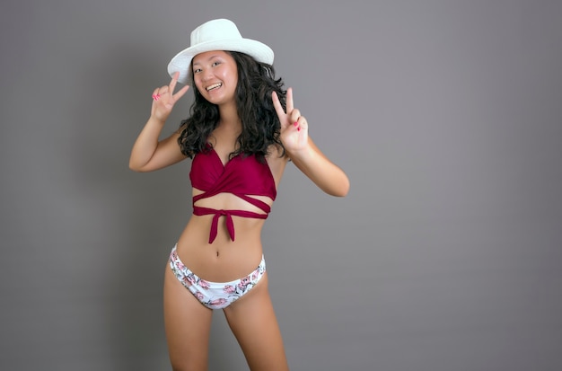 Hübsches junges chinesisches Mädchen im Bikini mit Hut, der lächelt und die Siegesgeste mit beiden Händen macht