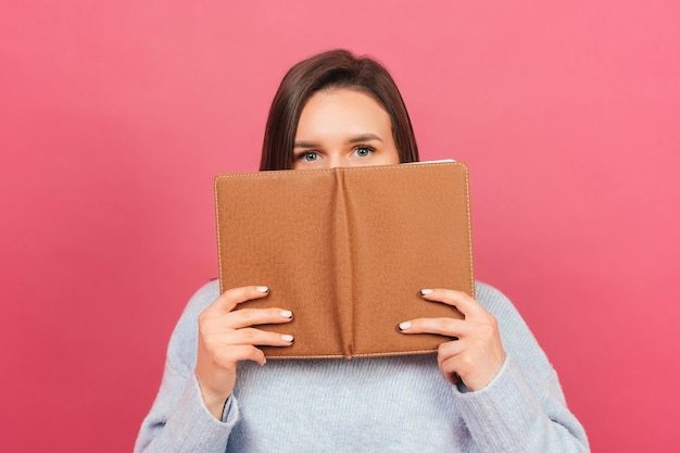 Hübsches junges Mädchen bedeckt das halbe Gesicht mit einem braunen Notizbuch