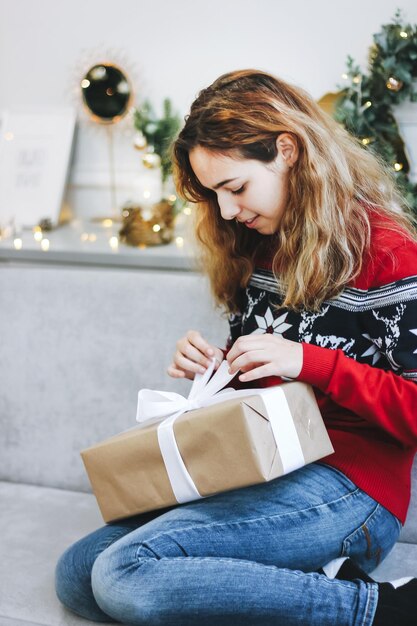 Hübsches junges Mädchen, das Geschenke nahe Weihnachtsbaum öffnet. Mädchen in einem roten Strickpullover hält Geschenkbox.