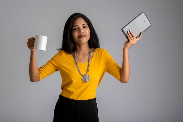 Hübsches junges Mädchen mit einer Tasse Tee oder Kaffee unter Verwendung des Smartphones, das auf graue Wand aufwirft.