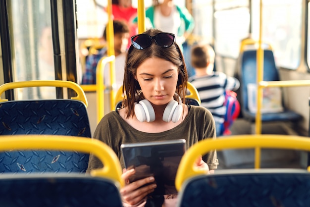 Hübsches junges Mädchen mit Kopfhörern, die in einem Bus sitzen und Tablette beobachten.