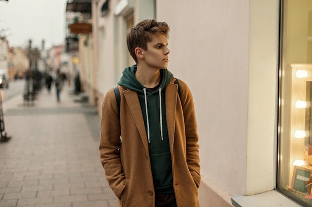 Hübsches junges Modell eines Mannes in einem trendigen Mantel mit einem Kapuzenpulli geht in der Stadt nahe dem Schaufenster spazieren
