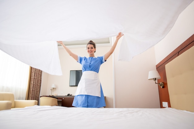 Hübsches junges Zimmermädchen in Uniform, das weiße saubere Wäsche über das Bett streckt, während es im Hotelzimmer arbeitet