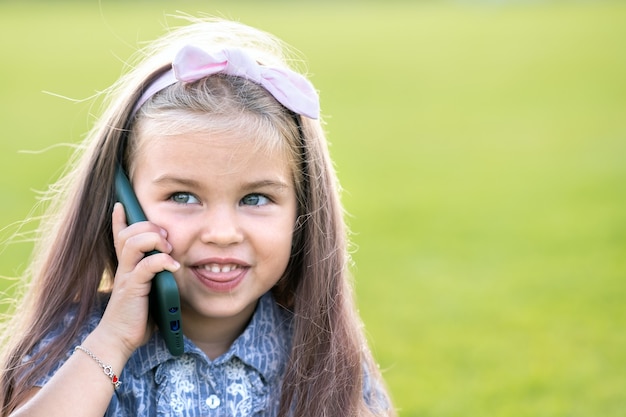 Hübsches kleines Kindermädchen, das im Sommer glücklich im Freien lächelt und am Handy spricht.