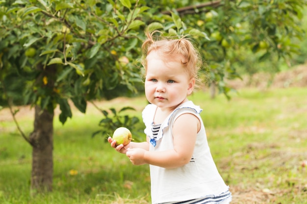 Hübsches kleines Mädchen, das einen Apfel in der Hand hält und unter dem Apfelbaum steht