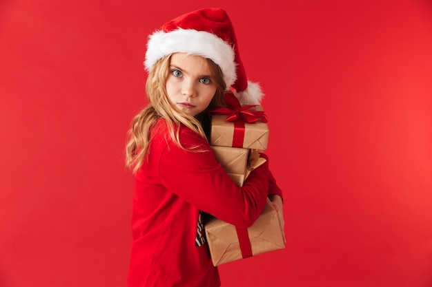 Hübsches kleines Mädchen, das Weihnachtsmütze trägt, isoliert steht und Stapel von Geschenkboxen hält