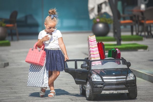 Hübsches kleines Mädchen Fashionista, das Einkaufen mit Einkaufstaschen macht. Nettes Kind draußen im Spielzeugauto