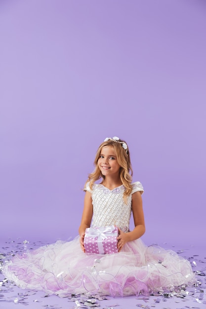 Hübsches kleines Mädchen gekleidet in Prinzessinnenkleid, das isoliert über violetter Wand sitzt und Geschenkbox hält