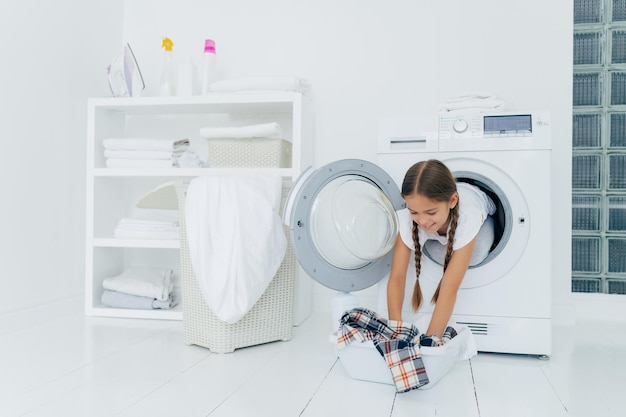 Hübsches kleines Mädchen Haushaltsvorstand posiert in der Waschmaschine nimmt kariertes Hemd aus dem Becken, das mit Wäsche beschäftigt ist, hat einen frohen Ausdruck, zwei gekämmte Zöpfe Kindheit und Waschtag Konzept