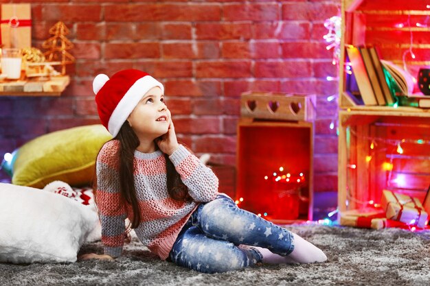 Hübsches kleines Mädchen im roten Hut, das im weihnachtlich dekorierten Raum sitzt