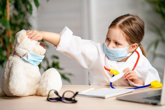 Hübsches kleines Mädchen in einem weißen Laborkittel und einer Maske nahm die Temperatur eines Teddybären an.