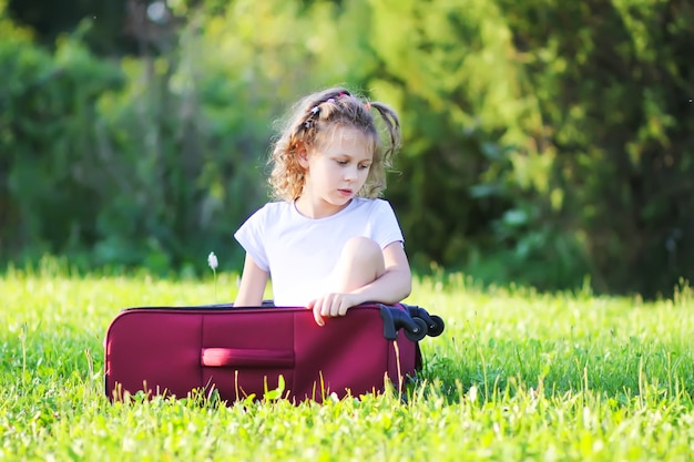Hübsches kleines Mädchen mit dem lockigen Haar, das draußen in der Reisetasche auf der grünen Sommerwiese sitzt