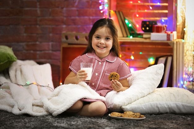 Hübsches kleines Mädchen mit Glas Milch und leckeren Keksen im weihnachtlich dekorierten Raum