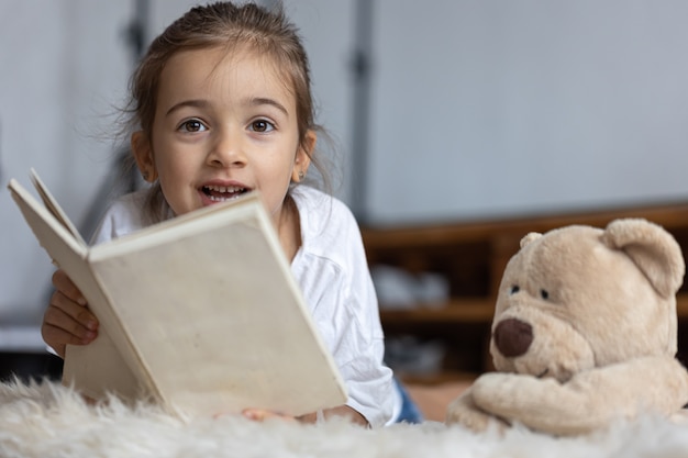 Hübsches kleines Mädchen zu Hause, das mit ihrem Lieblingsspielzeug auf dem Boden liegt und Buch liest.