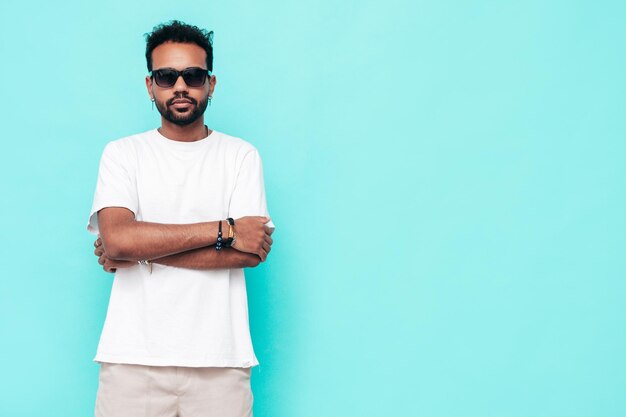 Hübsches lächelndes Hipster-Model Sexy unrasierter Mann in weißem Sommert-shirt und Jeanskleidung Modemann posiert in der Nähe der blauen Wand im Studio Isoliert mit Sonnenbrille