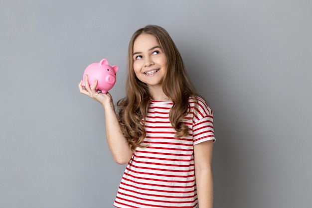 Hübsches lächelndes kleines Mädchen, das ein T-Shirt trägt, das eine Schweinespardose in den Händen hält, um Geld beim Banking zu sparen