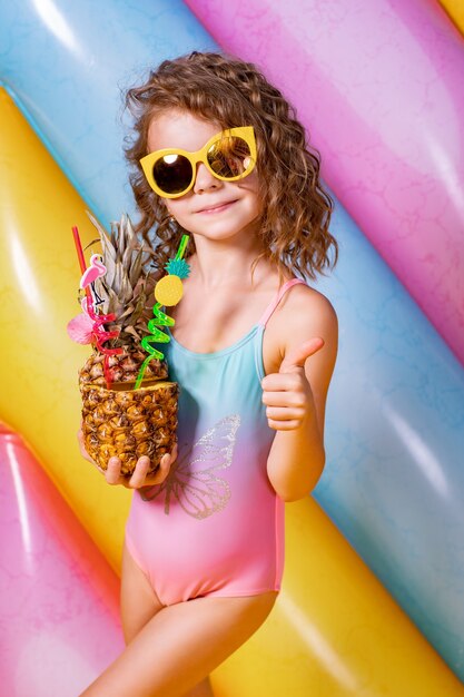 Hübsches lächelndes Mädchen, das rosa und blaue Badebekleidung und Sonnenbrille hält, die Ananas-Cocktail mit bunten Strohhalmen auf aufblasbarem Regenbogenmatratzenraum halten