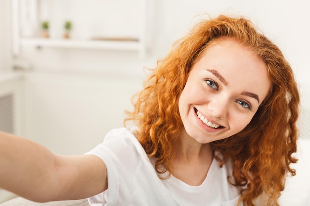 Hübsches lächelndes Mädchen, das selfie Nahaufnahme macht. Glücklicher Rotschopf in weißer Freizeitkleidung drinnen.