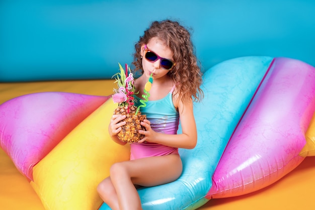 Hübsches lächelndes Mädchen, welches die rosa und blaue Badebekleidung und Sonnenbrille hält Ananascocktail mit bunten Strohen trägt und Daumen oben auf aufblasbarer Matratze des Regenbogens zeigt