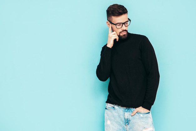 Hübsches lächelndes Modell Sexy stilvoller Mann in schwarzem Rollkragenpullover und Jeans gekleidet Mode-Hipster-Männchen posiert in der Nähe der blauen Wand im Studio Isoliert