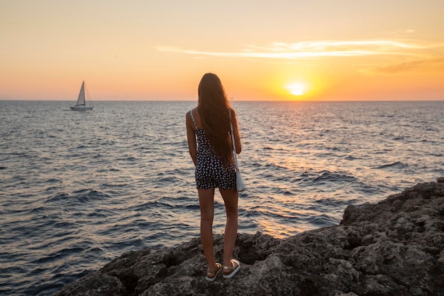 Hübsches Mädchen blickt bei Sonnenuntergang auf den Ozean, gekleidet in Sommerkleidung