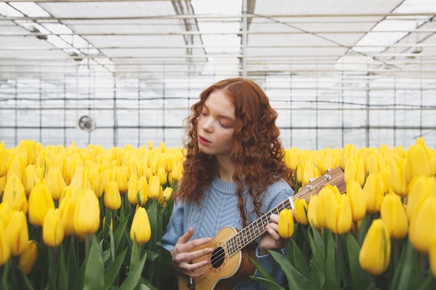 Hübsches Mädchen, das Gitarre spielt, während es zwischen gelben Tulpen im Gewächshaus steht