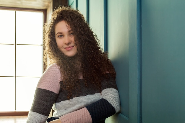 Hübsches Mädchen, das nahe der Wand im Hintergrundfenster steht. Locken von langen Haaren. Winterpullover.