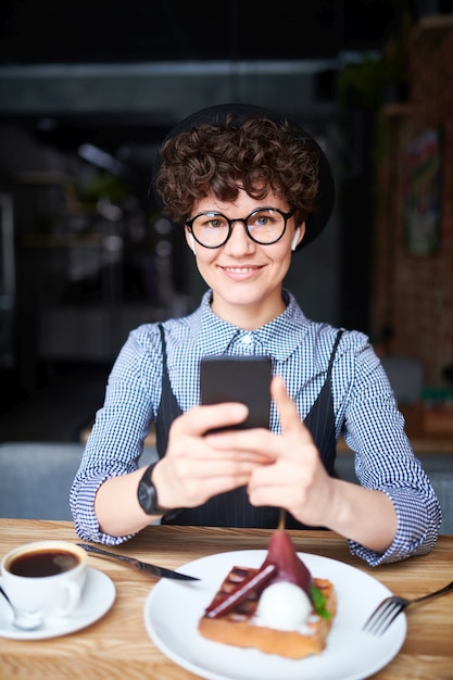 Hübsches Mädchen im Hut und im intelligenten lässigen Scrollen in ihrem Smartphone, während im Café mit Getränk und Nachtisch in der Nähe sitzen