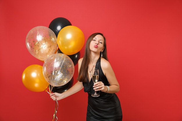 Hübsches Mädchen im schwarzen Kleid, das blasende Lippen feiert, senden Luftkuss halten Glas Champagner-Luftballons einzeln auf rotem Hintergrund. Valentinstag Happy New Year Geburtstag Mockup Urlaub Party Konzept.