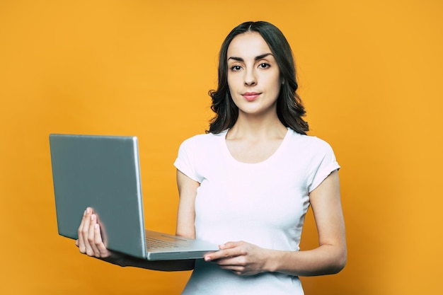 Hübsches Mädchen im weißen Basic-T-Shirt bleibt vor orangefarbenem Hintergrund und hält einen grau-silbernen Laptop in den Händen