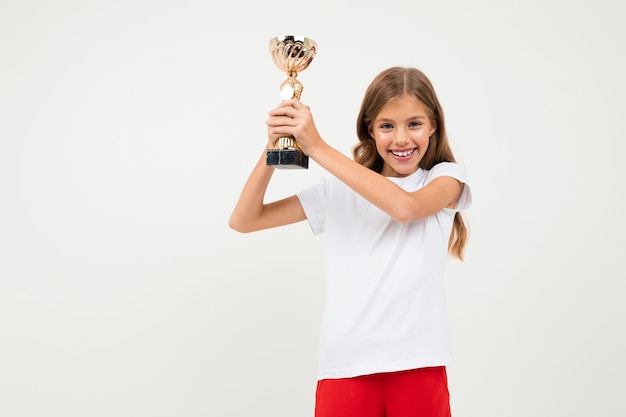 Hübsches Mädchen im weißen und roten Kostüm zeigt ihre goldene Tasse und freut sich auf weißer Wand