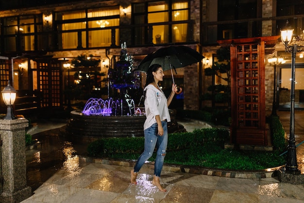 Hübsches Mädchen in der regnerischen Nacht im Freien Porträt einer schönen Frau, die mit Regenschirm unter Regen steht, gute Laune bei kaltem, nassem Wetter