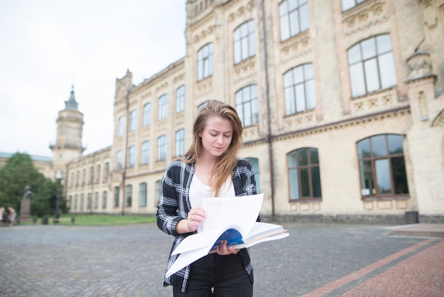 Hübsches Mädchen in Freizeitkleidung steht auf dem Campus und liest ein Buch. Studieren an der Universität.