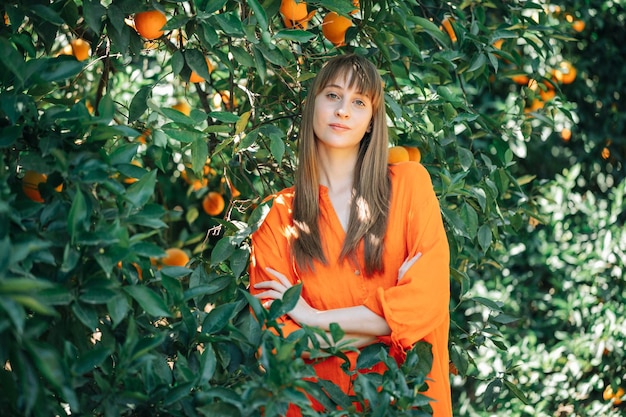 Hübsches Mädchen in orangefarbenem Kleid posiert vor der Kamera, indem es die Arme im Orangengarten kreuzt